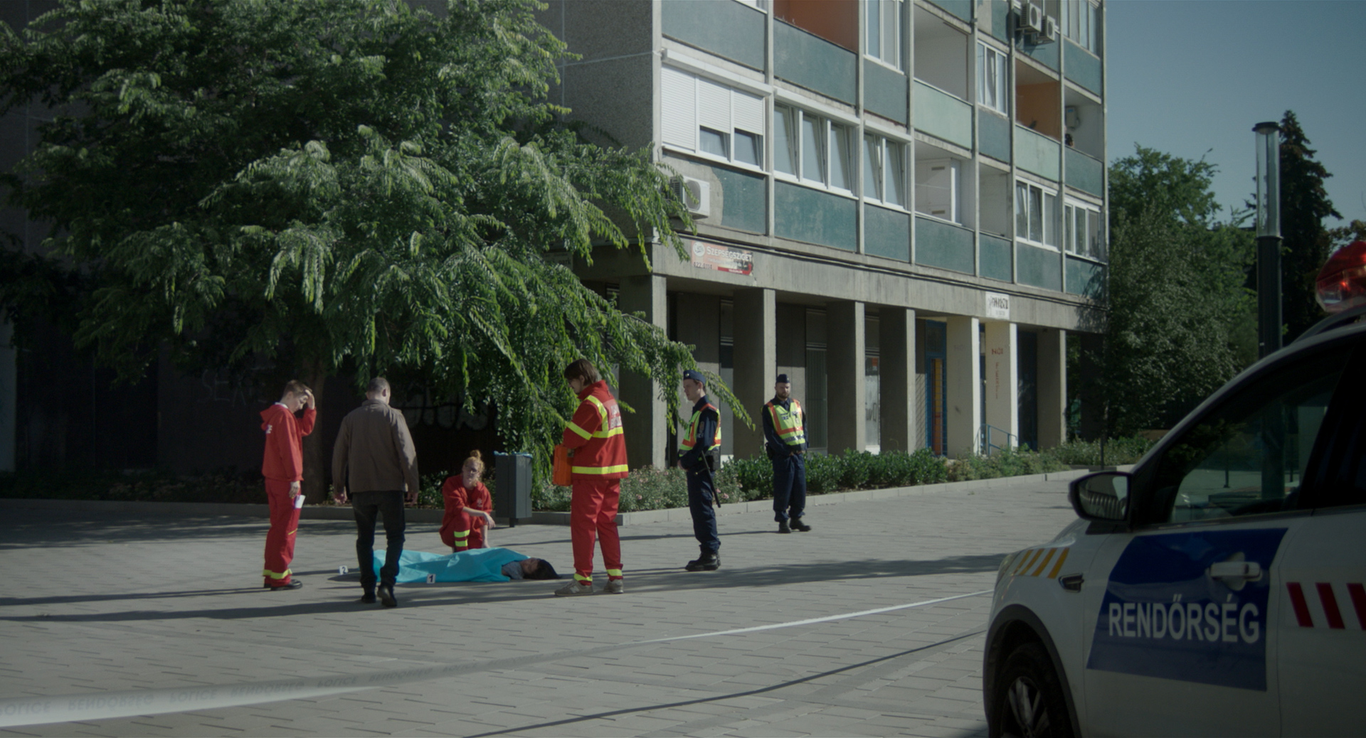 eszter galambos, Still from the short Tiger. Director of Photography, DOP, Cinematographer, operatőr: Eszter Galambos. Director: Péter Hajmási. Colorist: Anna Stalter, Cast: Milán SchruffA dead chinese women is lying on the ground. Medics and the police are looking at her. She is covered with a blue blanket. Police car stops. Still from the movie short Tiger. DOP. : Eszter Galambos  